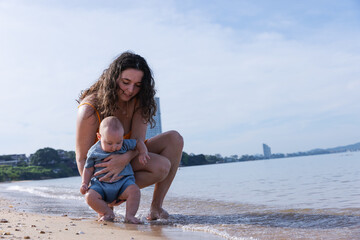Fototapeta premium Mother Gently Holding Baby by the Shoreline, Introducing Her Child to the Ocean Waves in a Relaxed Beach Environment, Sharing a Precious Moment in Nature on a Calm, Sunny Day
