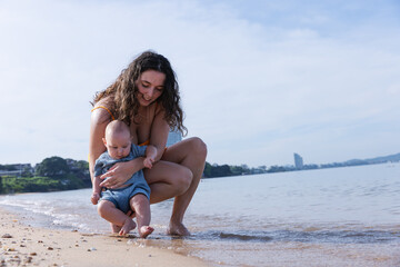Naklejka premium Mother Gently Holding Baby by the Shoreline, Introducing Her Child to the Ocean Waves in a Relaxed Beach Environment, Sharing a Precious Moment in Nature on a Calm, Sunny Day