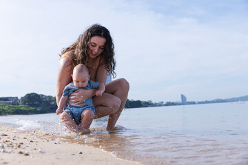 Fototapeta premium Mother Gently Holding Baby by the Shoreline, Introducing Her Child to the Ocean Waves in a Relaxed Beach Environment, Sharing a Precious Moment in Nature on a Calm, Sunny Day