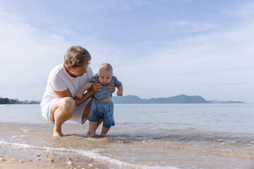 A loving grandmother helps her baby grandchild explore the shoreline, gently dipping the baby's...