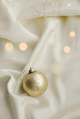Gold Christmas Ornament on Soft White Fabric. Christmas decoration featuring a glittering golden bauble resting on a smooth white fabric background with soft, warm fairy lights, minimalism and winter-