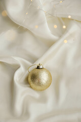 Gold Christmas Ornament on Soft White Fabric. Christmas decoration featuring a glittering golden bauble resting on a smooth white fabric background with soft, warm fairy lights, minimalism and winter-