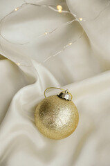 Gold Christmas Ornament on Soft White Fabric. Christmas decoration featuring a glittering golden bauble resting on a smooth white fabric background with soft, warm fairy lights, minimalism and winter-