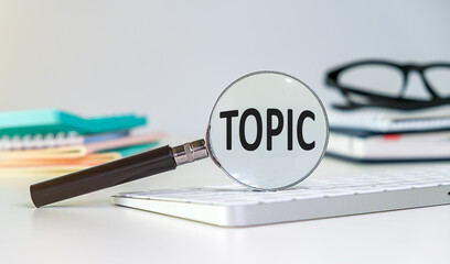 The word TOPIC is written on a magnifying glass on office background.