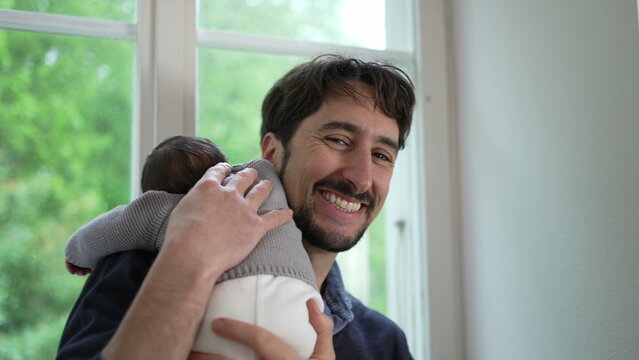 Newborn baby resting on the father’s shoulder, dressed in a cozy knitted sweater. The father turns body towards window, highlighting the comforting and secure embrace between them