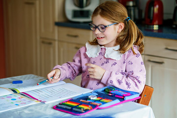 Little girl with glasses, wearing a pink sweater, sits at a table doing homework. Child looks up with a smile, holding a pencil, with an open book in front of her. Indoors, studying, and engaged.