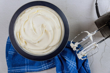 Large bowl filled with fluffy whipped cream sits on countertop, accompanied by mixer on blue towel. It looks fresh and ready for use in desserts or toppings.