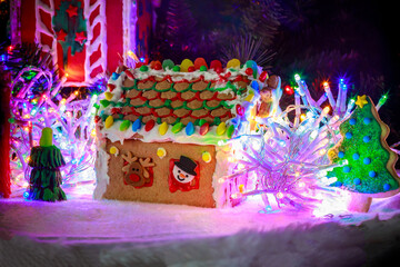 Gingerbread house with bright, colorful roof made of candy and icing sits amidst glowing lights and decorative tree during holiday season.