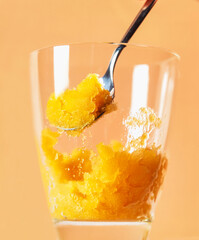 Refreshing Mango Sorbet Served In A Clear Glass With A Spoon. A bright yellow mango sorbet is scooped from a glass, illuminated against a warm orange background