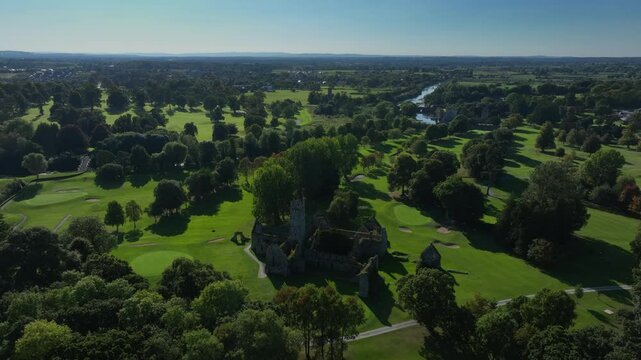 Franciscan Friary, Adare, County Limerick, Ireland, September 2024. Drone orbits clockwise while descending towards the building behind trees on the bright side of ruins in middle of a golf course.