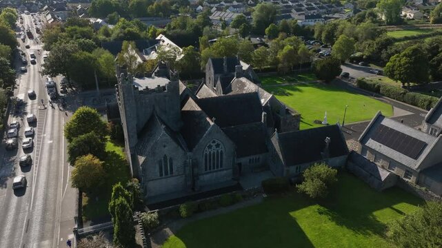 Holy Trinity Abbey Church, Adare, County Limerick, Ireland, September 2024. Drone orbits clockwise from shaded side of church by the main street as light glistens and shines off adjacent rooftops.