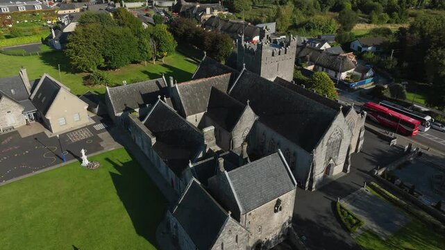 Holy Trinity Abbey Church, Adare, County Limerick, Ireland, September 2024. Drone orbits clockwise above angled slate roofs of church building with school playground and grassy field behind.
