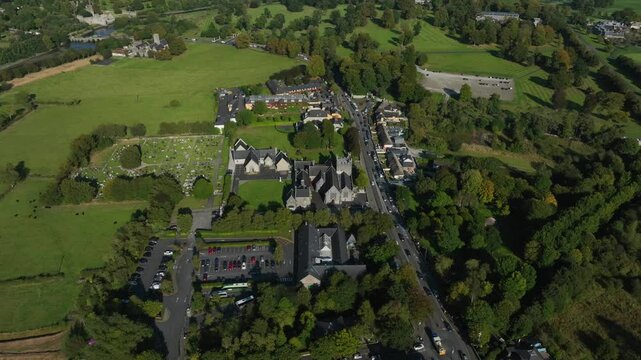Holy Trinity Abbey Church, Adare, County Limerick, Ireland, September 2024. Drone orbits clockwise and descends over center street of cars driving through town with cemetery and open fields behind.