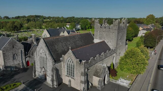Holy Trinity Abbey Church, Adare, County Limerick, Ireland, September 2024. Drone orbits clockwise in a medium close up of the gothic stone building with stained glass windows rising above tree line.