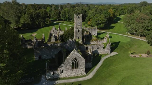 Franciscan Friary, Adare, County Limerick, Ireland, September 2024. Drone orbits counter clockwise and rises behind a tree revealing the illuminated broken walls and towers of decaying old building.
