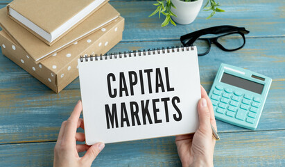 Businessman holding a card with text CAPITAL MARKETS. Wooden background