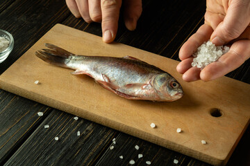 Hands sprinkle salt over a whole fish laid on a rustic wooden board. The kitchen exudes warmth, with a focus on the process of seasoning the fresh catch, ready for cooking.