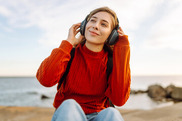 Relaxed woman enjoying music by the waterfront while wearing headphones by a lighthouse. Travel, tourism, technology, blogging concept.
