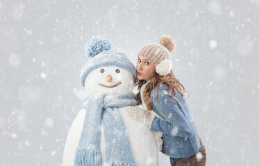 Happy young woman enjoying kissing a snowman in a winter snowfall, dressed in warm winter clothing. Ski trip during the Christmas holidays in winter season. Cooling and Heating temperatures Concept