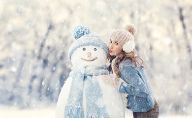 Happy young woman enjoying kissing a snowman in a winter snowfall, dressed in warm winter clothing. Ski trip during the Christmas holidays in winter season. Cooling and Heating temperatures Concept