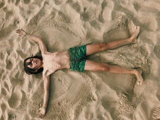 A young boy is comfortably resting on a sandy beach background, with his arms playfully outstretched