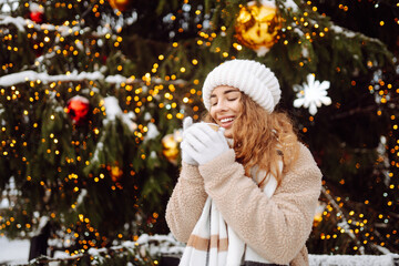 Happy woman drinks a hot drink at a Christmas market decorated with festive lights. Winter holidays, vacation, tourism concept.