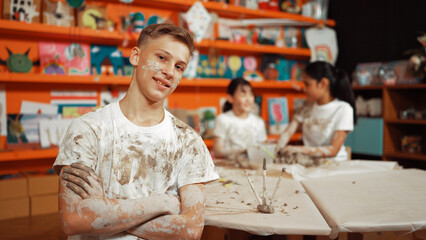 Smiling boy looking at camera and crossing arm with confident at workshop while diverse student having pottery class together. Happy caucasian student smile while pose with arm folded. Edification.