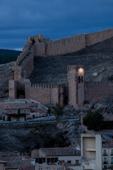Illuminated medieval watchtower and stone walls at dusk with rustic village houses below © larrui