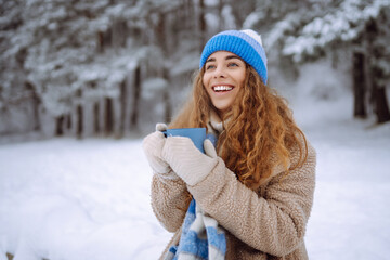 Pretty woman in winter hat enjoys the snow and drinks hot beverage standing outside on the snow in the forest. ravel, tourism, nature, active lifestyle.