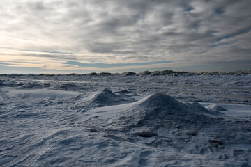 Snow-covered seashore, little snowy hills and hummocks of broken ice at the water's edge. Beautiful low cloudy sky and diffused yellow sunlight at the end of a short winter day