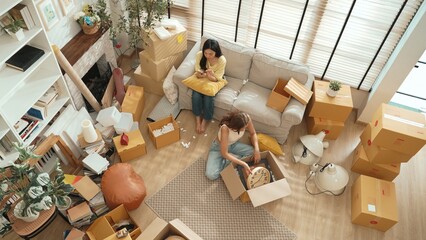 Top down view of friend unpacking box while roommate playing smart phone on sofa. Attractive woman checking furniture at package and preparing for moving while the other using social media. Myrmidon.