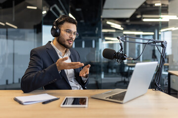 Man in suit with headphones speaks into microphone during podcast in office. Uses laptop and tablet...