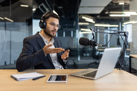 Man in suit with headphones speaks into microphone during podcast in office. Uses laptop and tablet while communicating effectively. Concept of technology, communication, and media.