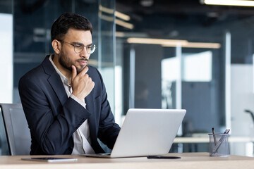 Businessman in dark suit analyzing work on laptop in office. He appears thoughtful and focused,...