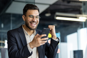 Excited businessman in suit celebrates success using smartphone in modern office. Expression of joy and accomplishment, highlighting business communication and digital connection.