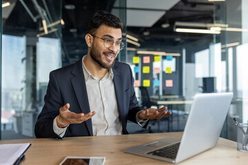 Businessman in modern office engaging in virtual meeting via laptop. Confident expression, open gestures highlight communication, professionalism. Smartphone on table indicates digital workspace.
