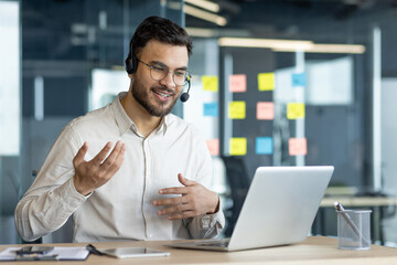 Business professional wearing headset in virtual meeting, engaging in remote conversation using laptop. Modern office setup highlights digital communication efficiency. Suitable for business context.