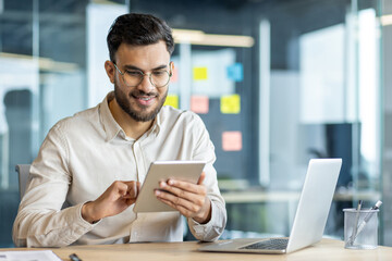 Smiling young professional using tablet in modern office. Male office worker multitasking,...