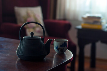 A cozy window lit indoor scene with cast iron teapot and a hand made ceramic teacup with floral design in front of a classic armchair, cushion and a coffee table 