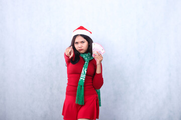 gesture of adult woman wearing red christmas sweater, santa hat and scarf, serious to camera hand holding rupiah cash money and pointing in front, you, isolated on white background