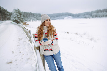 Fototapeta premium Happy woman drinks a hot drink from a thermos in a winter snowy forest. Warm up in frozen winter forest, warm hands. travel, wanderlus.