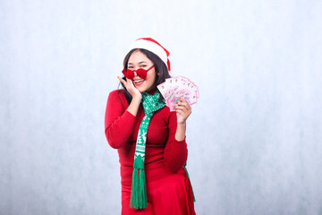 gesture of adult Asian woman wearing red christmas sweater, red glasses, Santa hat, and scarf, happy to camera hand holding rupiah cash money and cheek, isolated on white background