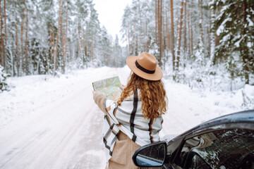Fototapeta premium Young woman with a map near the car on a snowy winter road in the forest. Woman on vacation looking at map for directions while driving in car.