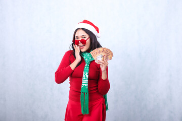 gesture of indonesia woman wearing red christmas sweater, red glasses, Santa hat and scarf, cheerful looking at camera hand holding euro cash money and beside mouth, isolated on white background