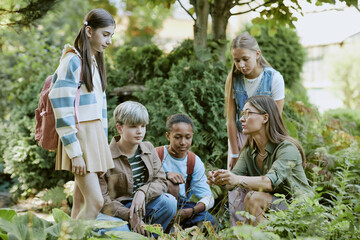 Young female botanist showing rare plants to group of teen students while visiting local botanical...