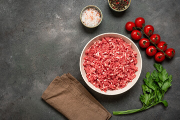 Raw mince beef, ground meat in bowl with spices and cherry tomatoes on dark grunge background, top view