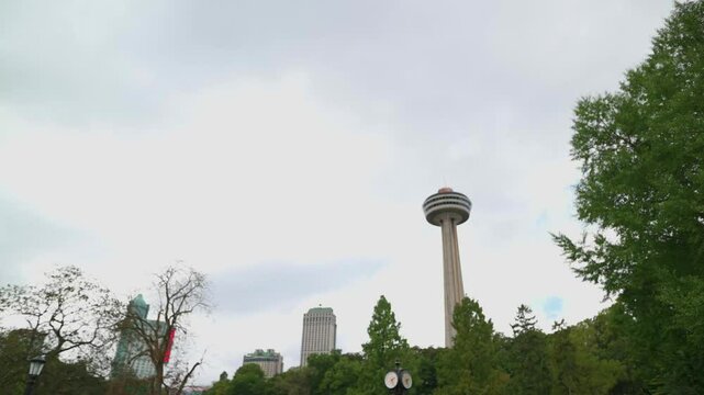 view of the Skylon Tower rising above trees and surrounding buildings under a cloudy sky, blending urban architecture with natural elements in Niagara Falls, Canada