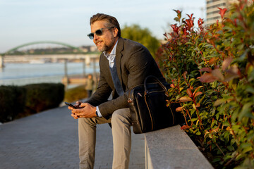 Obraz premium Man enjoying a peaceful evening by the riverside in casual elegance with a smartphone in hands and a scenic backdrop
