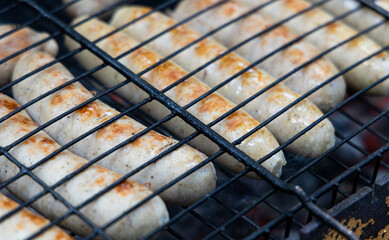 Close-up shot of juicy, golden brown sausages grilling on a black metal grill rack. The sausages are perfectly arranged, with visible grill marks, indicating they are nearing completion.