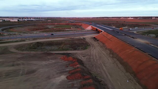 Low-altitude drone view from a newly paved highway overpass overlooking an active motorway during road construction. Modern transportation infrastructure with moving traffic, construction works and ex